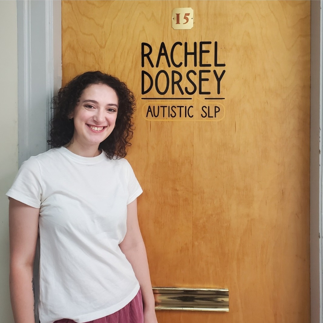 A photo of Rachel, a white woman with dark curly hair, standing in front of a door with her name on it.